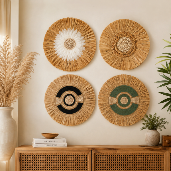Four round woven raffia wall hangings in natural tan, decorated with white, black, and green geometric and floral patterns, displayed on a beige wall above a wooden cabinet with pampas grass and plants.
